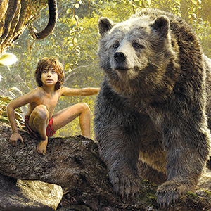 A young boy wearing a red loincloth sits on a rock in a jungle, touching the back of a large, friendly-looking bear standing beside him. Sunlight filters through the dense green foliage around them.