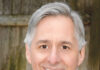 A middle-aged man with gray hair and a friendly smile, wearing a plaid shirt, poses in front of a wooden background.