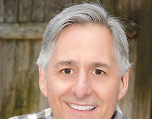 A middle-aged man with gray hair and a friendly smile, wearing a plaid shirt, poses in front of a wooden background.