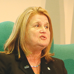 A middle-aged woman with shoulder-length blonde hair is speaking while seated in a teal chair. She is wearing a black blazer with pins and a beaded necklace. The background is softly lit and neutral-colored.