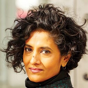 A person with short, curly black hair and a nose piercing looks at the camera with a slight smile, wearing a dark top and hoop earrings, with a blurred indoor background.
