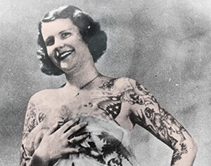 A vintage black-and-white photo of a smiling woman with short dark hair, covered in tattoos on her arms and chest, posing confidently with a sheer, patterned fabric draped around her torso.