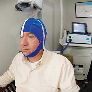 A man wearing a blue cap with white straps sits in a clinic while undergoing a medical procedure involving a machine with wires attached to his head.