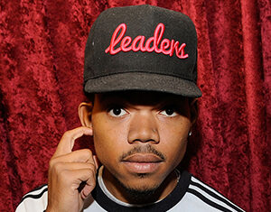 A man wearing a black cap with red cursive lettering and a light-colored sweatshirt stands in front of a red curtain, touching his ear and looking directly at the camera.