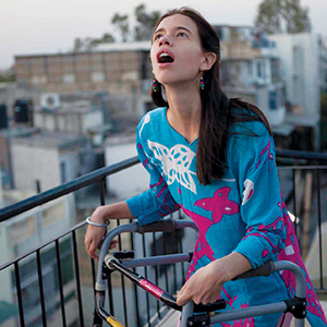 A woman with long dark hair, wearing a bright blue and pink top, stands on an outdoor balcony with a walker, looking up with an amazed expression. Urban buildings are visible in the background.