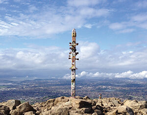 A tall, colorful totem pole stands among rocky terrain atop a hill, overlooking a sprawling city and landscape under a partly cloudy sky.