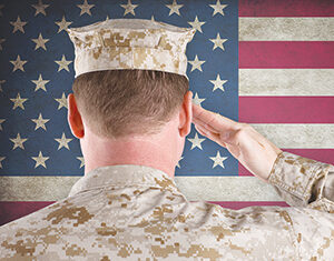 A person in military camouflage uniform salutes with their right hand, facing an American flag in the background. The view is from behind, showing the back of the uniform and the flag’s stars and stripes.