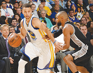 A basketball player in a white and yellow Golden State Warriors uniform dribbles past a defender in a black San Antonio Spurs uniform during a game, with spectators watching in the background.