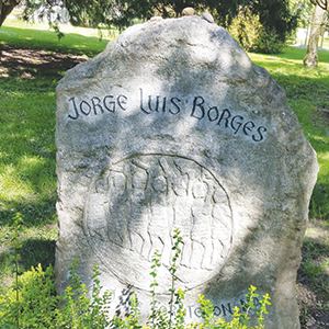 A large stone memorial in a grassy area, engraved with Jorge Luis Borges at the top and a circular design beneath the name. Sunlight filters through nearby trees onto the stone.