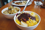 A small bowl of fruit and granola topped with yogurt and berry sauce sits in front of a bowl of roasted potatoes on a wooden table, with another plate blurred in the background.