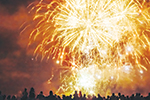 A large crowd watches bright yellow and orange fireworks exploding in the night sky, lighting up the clouds above.