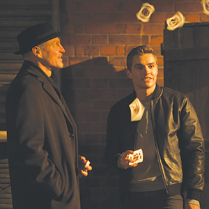 Two men stand outside at night near a brick wall. One wears a dark coat and hat, while the other, in a black jacket, fans out playing cards, some of which are mid-air. The scene is dimly lit.