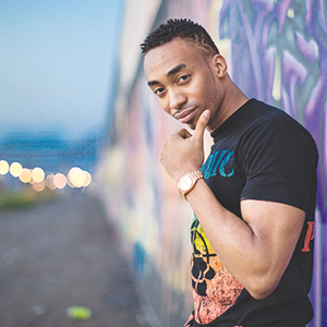 A man wearing a black t-shirt and gold watch stands outdoors by a graffiti-covered wall, looking at the camera with a slight smile and his hand resting on his chin. The background is blurred with city lights.