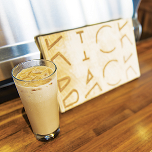 A glass of iced coffee sits on a wooden countertop next to a wooden sign with the words KICK BACK carved into it.