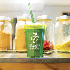 A green smoothie in a plastic cup with a straw and a yellow lid sits on a counter. Behind it are glass jars filled with various powders and dried ingredients. The cup has a logo that says vitamina juice & blends.