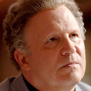 A middle-aged man with curly light brown hair looks slightly upward with a thoughtful expression, wearing a gray jacket and a dark shirt, against a softly blurred background.