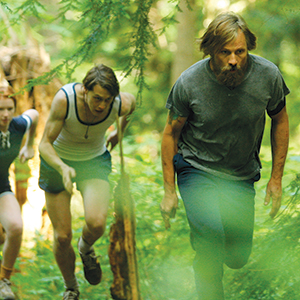 Three people, including a bearded man in a gray shirt, are running through a lush, green forest. Two others, wearing athletic clothes, are closely following him along a wooded trail. Sunlight filters through the trees.