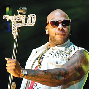 A man wearing sunglasses and a sleeveless studded vest holds a microphone stand with a jeweled sign spelling FLO shaped like Florida, performing onstage under bright lights.