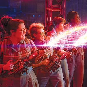 Four women in jumpsuits use glowing, high-powered proton blasters, emitting colorful energy beams, in a dramatic, action-packed scene with vibrant red and purple lighting.