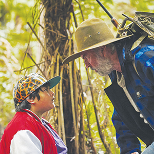 An older man with a beard and wide-brimmed hat leans toward a young boy in a red jacket and animal-print cap, both standing in a forest and gazing seriously at each other.