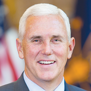 A middle-aged man with short white hair is smiling in a formal portrait. He is wearing a dark suit and a light-colored shirt. The blurred background includes an American flag and other indistinct details.