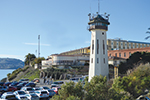A historic prison building with a tall guard tower overlooks a parking lot filled with cars; hills and water are visible in the background under a clear sky.