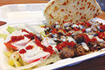 A takeout container holding gyro meat topped with white and red sauces, sliced tomatoes with lettuce, and a piece of folded flatbread.