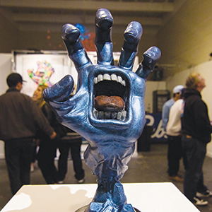 A blue sculpture of a hand with an open mouth in its palm, showing teeth and tongue, is displayed on a pedestal. Several people are gathered in the background at an art exhibit.