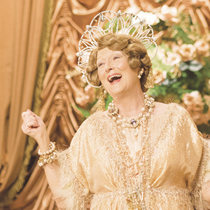 An older woman in an elaborate gold dress and ornate headpiece smiles and gestures joyfully while standing in front of gold drapes and floral decorations.
