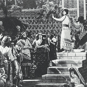 A black-and-white scene shows a woman in ancient-style clothing standing on steps, surrounded by people in robes. The background features ornate patterns and palm trees, evoking a historical or theatrical setting.