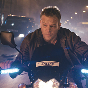 A man in a leather jacket rides a police motorcycle at night, with blue lights flashing. The word “POLICE” and Greek text are visible on the bike’s front, and a police van is blurred in the background.