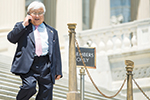 An older man in a suit and tie talks on a cellphone while walking down steps outside a government building. A sign reading Members Only hangs on a rope nearby.