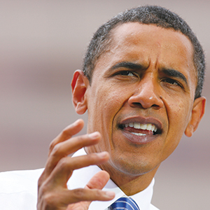 A man in a white shirt and blue-striped tie gestures with his hand while speaking, appearing focused and expressive. The background is blurred.