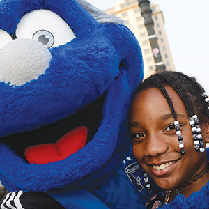 A person with beaded braids smiles next to a large blue mascot with a big nose, wide eyes, and an open mouth, outdoors with a building in the background.