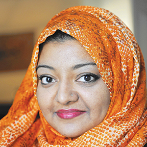 A woman wearing an orange patterned hijab and pink lipstick looks at the camera with a slight smile. The background is softly blurred.