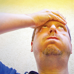 A man holds his forehead with one hand and looks upward with a concerned or stressed expression, standing against a light-colored wall with warm light at the top of the image.