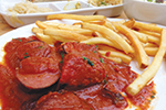 Sliced sausage covered in tomato-based sauce served with French fries on a white plate, with side dishes in the background.
