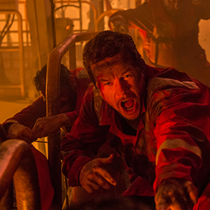 A man in a red jumpsuit, covered in dirt and blood, yells in distress while reaching forward. The scene is chaotic and dimly lit, suggesting an emergency or disaster situation with other figures in the background.