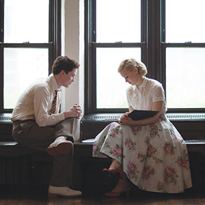 A young man and woman sit facing each other on a windowsill. The man wears a white shirt and tie, while the woman wears a white blouse and a floral skirt. They look down, appearing to have a quiet, thoughtful conversation.