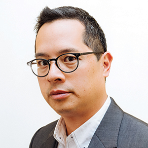 A man with short black hair and glasses wearing a white collared shirt and a gray suit jacket, looking slightly to the side against a plain white background.