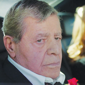 An elderly man with gray hair, dressed in a black suit and white shirt, sits in a car and looks solemnly out the window.