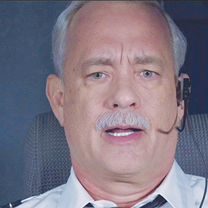 A middle-aged man with gray hair and a mustache wears a pilot uniform and a headset, sitting in what appears to be an airplane cockpit, looking forward with a serious expression.