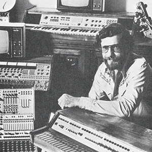 A man with glasses and a beard sits among vintage electronic music equipment, including synthesizers, control panels, and screens, in a black-and-white photo.