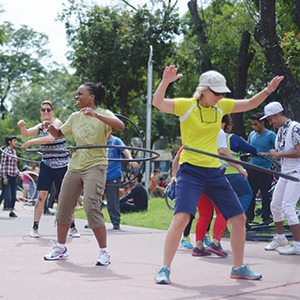 A group of people outdoors smiling and spinning hula hoops around their waists, with trees and more people in the background, suggesting a fun, active event in a park.