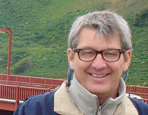 A man with short gray hair and glasses, wearing a beige and blue jacket, smiles in front of a red bridge with green hills in the background.
