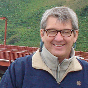 A man with short gray hair and glasses, wearing a beige and blue jacket, smiles in front of a red bridge with green hills in the background.