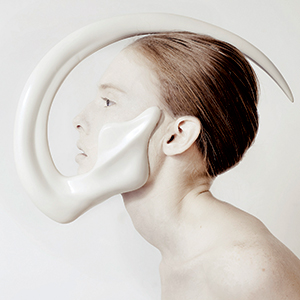 A person with pale skin and slicked-back hair wears a futuristic, sculptural white mask that curves dramatically from their jawline up over their head, set against a plain background.