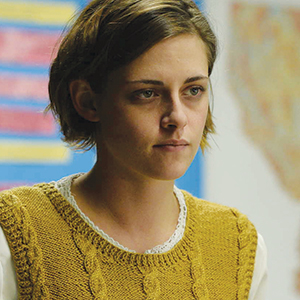 A person with short brown hair wearing a mustard yellow sweater over a white shirt sits indoors, looking slightly to the side. The background is colorful and blurred.