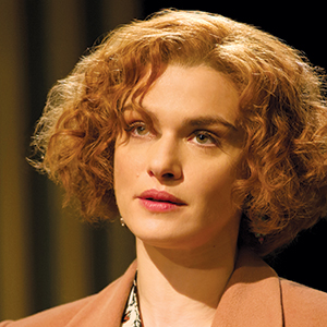 A woman with short, curly red hair is wearing a light brown coat and looking slightly to the side with a neutral expression, standing against a dark, blurred background.