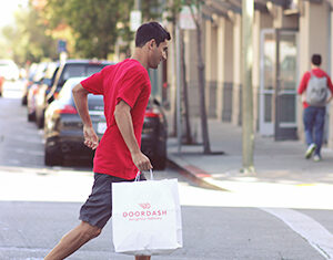 A man in a red shirt and blue sneakers crosses a street carrying a white DoorDash delivery bag. Cars and a building line the street, and another person with a backpack walks in the background.